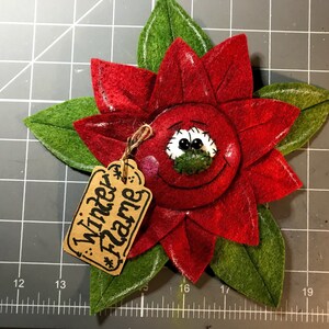 May include: A handmade felt poinsettia flower with a smiling face and green leaves. The red flower has a tag that reads "Winter Flame". The poinsettia is a festive holiday decoration.