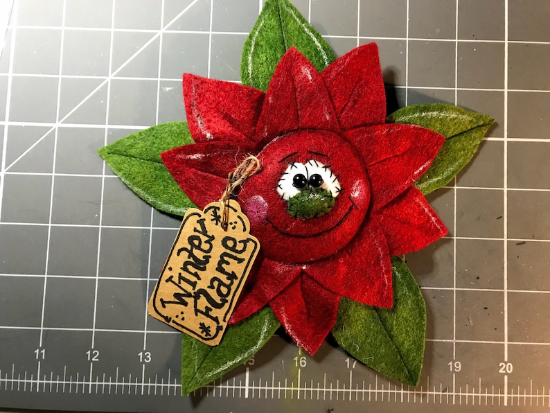 May include: A handmade felt poinsettia flower with a smiling face and green leaves. The red flower has a tag that reads "Winter Flame". The poinsettia is a festive holiday decoration.