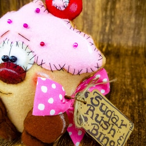 May include: A pink and brown felt cupcake with a red cherry on top and a pink and white polka dot bow. The cupcake has a brown felt base and a tag that says "Sugar Rush Bing".