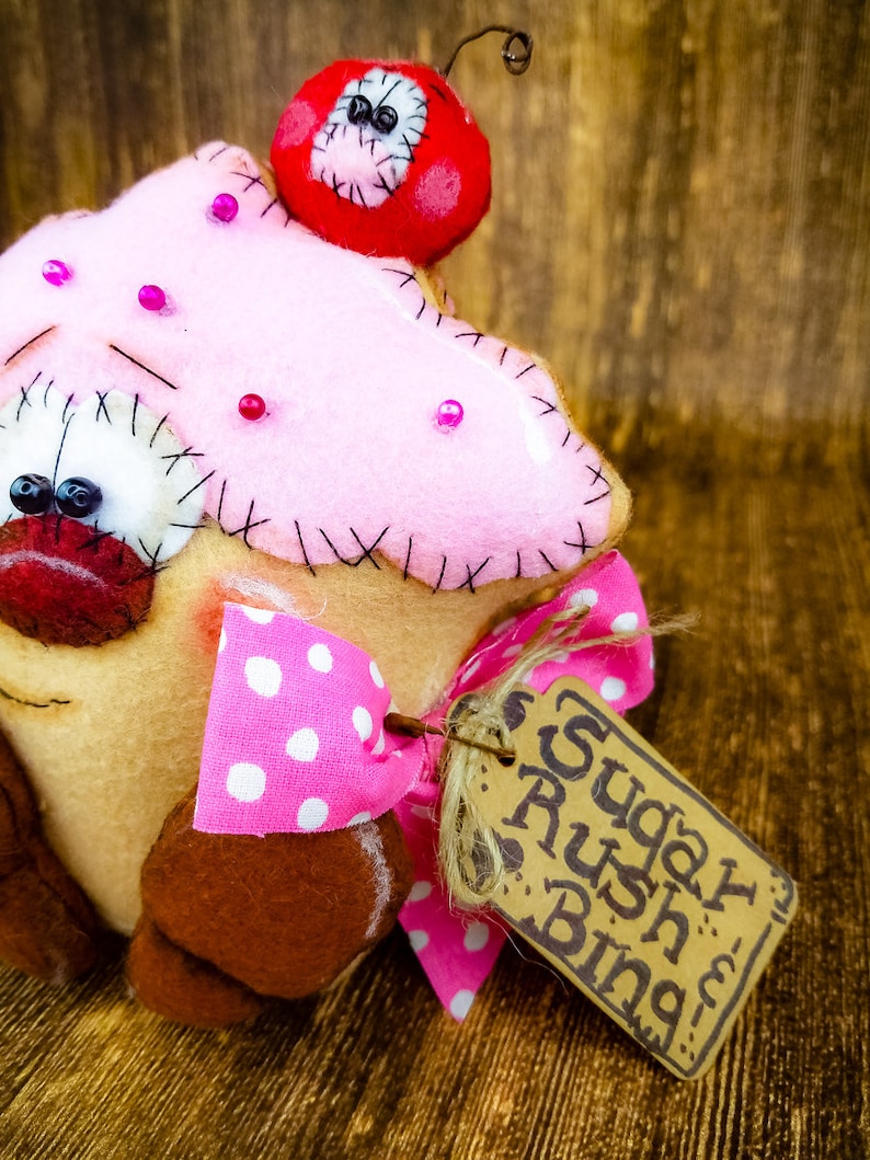 May include: A pink and brown felt cupcake with a red cherry on top and a pink and white polka dot bow. The cupcake has a brown felt base and a tag that says "Sugar Rush Bing".
