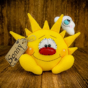 May include: A yellow felt sun with a stitched-on smile and a red nose. The sun has a white cloud with a blue eye on top of it. A brown tag with the word "Sonny" is attached to the sun.