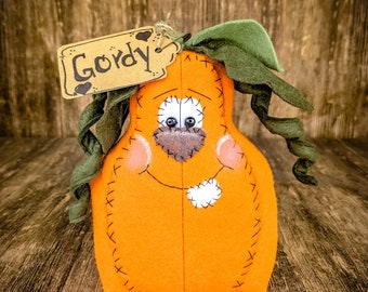 May include: A handmade orange pumpkin with a stitched smile and a green felt stem. The pumpkin has a brown felt nose and black eyes. A brown tag with the word "Gordy" is attached to the stem.