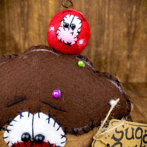 May include: A brown felt doll with a red mouth, black eyes, and white stitching around the eyes. The doll has a red felt hat with white stitching and black eyes. The doll is holding a brown paper tag with the text "Sugar Rus & Pie".