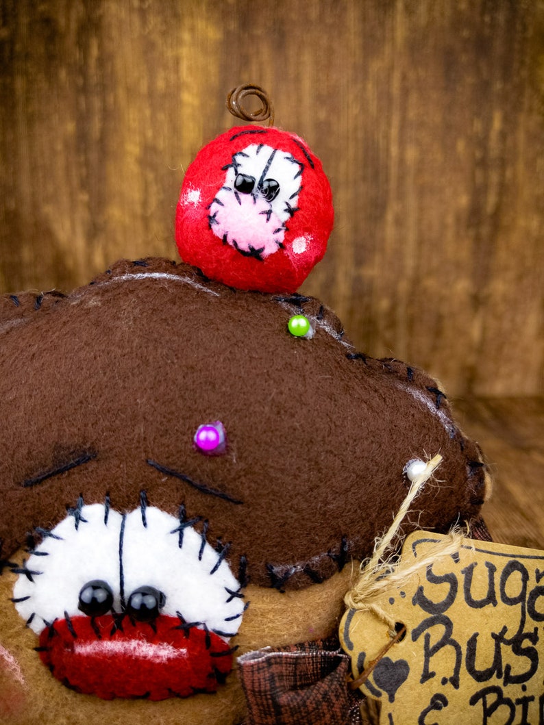 May include: A brown felt doll with a red mouth, black eyes, and white stitching around the eyes. The doll has a red felt hat with white stitching and black eyes. The doll is holding a brown paper tag with the text "Sugar Rus & Pie".