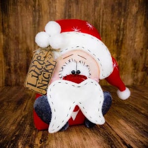 May include: A plush Santa Claus doll with a red hat featuring white pom-poms and white star accents. The doll has a pale face, black stitched eyes, a red nose, and a white felt moustache. A brown paper tag attached to the hat reads Little Nicky.