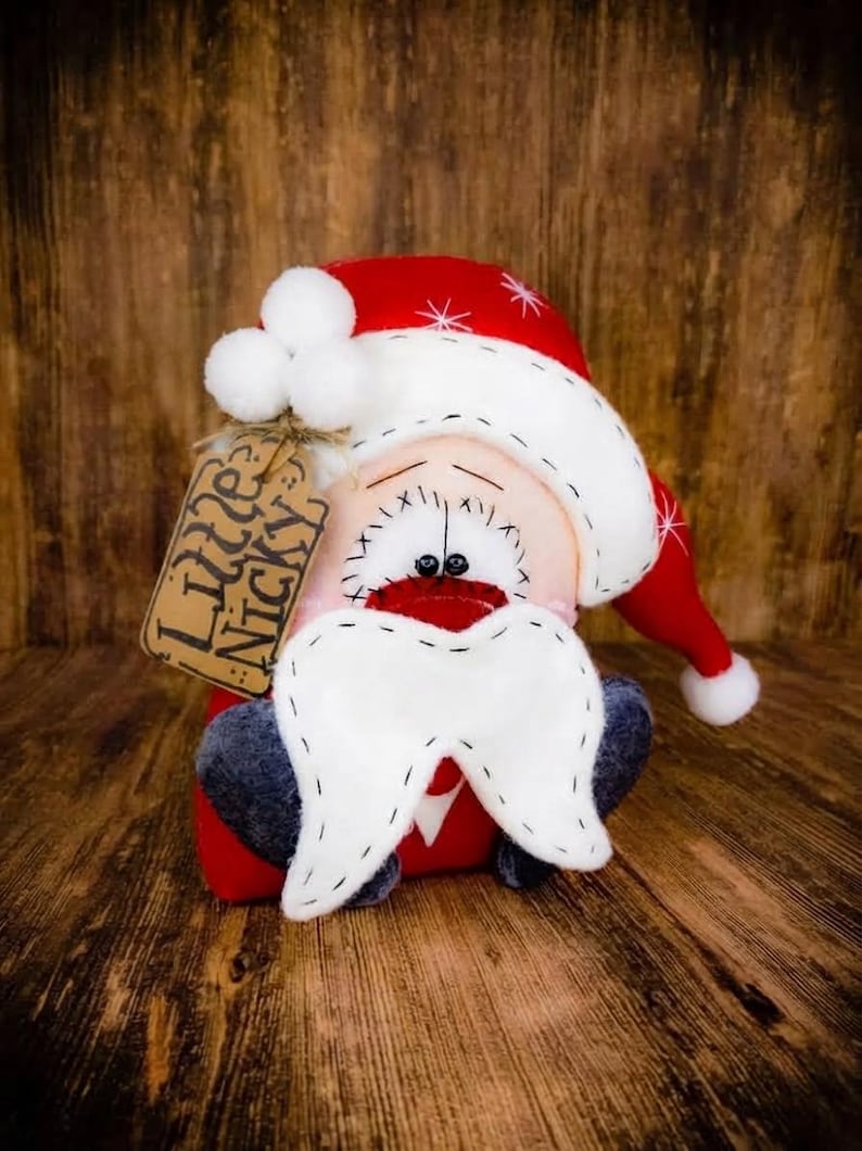 May include: A plush Santa Claus doll with a red hat featuring white pom-poms and white star accents. The doll has a pale face, black stitched eyes, a red nose, and a white felt moustache. A brown paper tag attached to the hat reads Little Nicky.