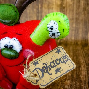 May include: A red felt apple with a green felt worm coming out of the side. The apple has a stitched-on face with googly eyes and a brown felt stem. A brown paper tag with the word "Delicious" written in black is attached to the apple.