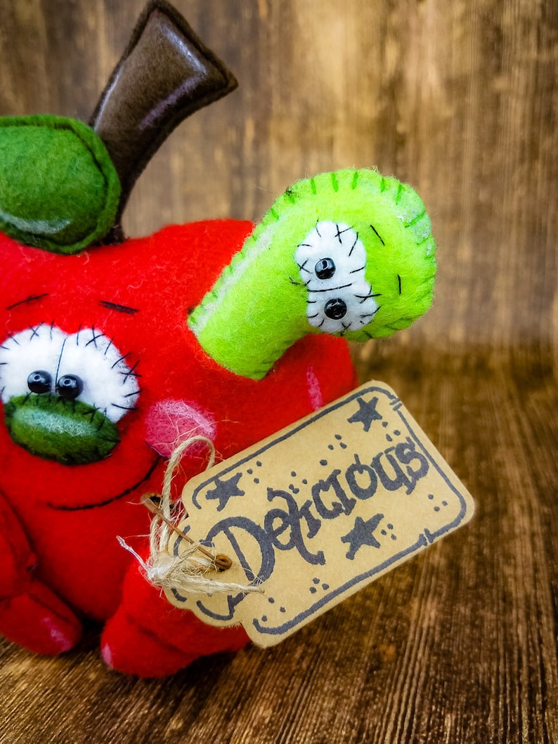 May include: A red felt apple with a green felt worm coming out of the side. The apple has a stitched-on face with googly eyes and a brown felt stem. A brown paper tag with the word "Delicious" written in black is attached to the apple.