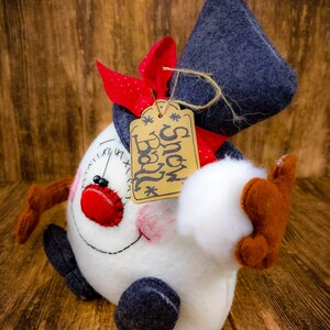 May include: A white snowman with a red nose and black eyes, wearing a blue hat and a red and white scarf. The snowman is holding a white snowball in its right hand. A brown tag with the text "Snow Ball" is attached to the hat.