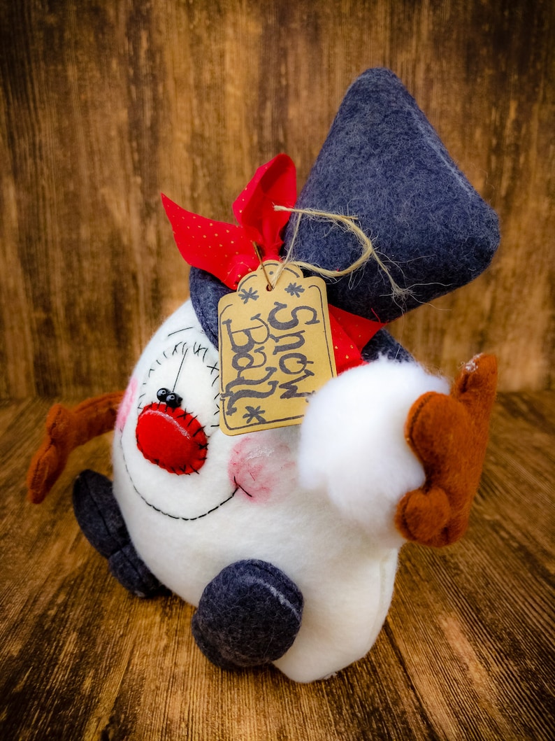 May include: A white snowman with a red nose and black eyes, wearing a blue hat and a red and white scarf. The snowman is holding a white snowball in its right hand. A brown tag with the text "Snow Ball" is attached to the hat.