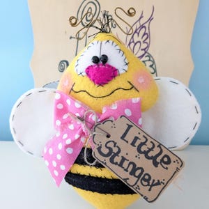 May include: A yellow felt bee with black stripes, white wings, and a pink bow. The bee has a friendly face with big eyes and a pink nose. It has a brown tag that says "Little Stinger".