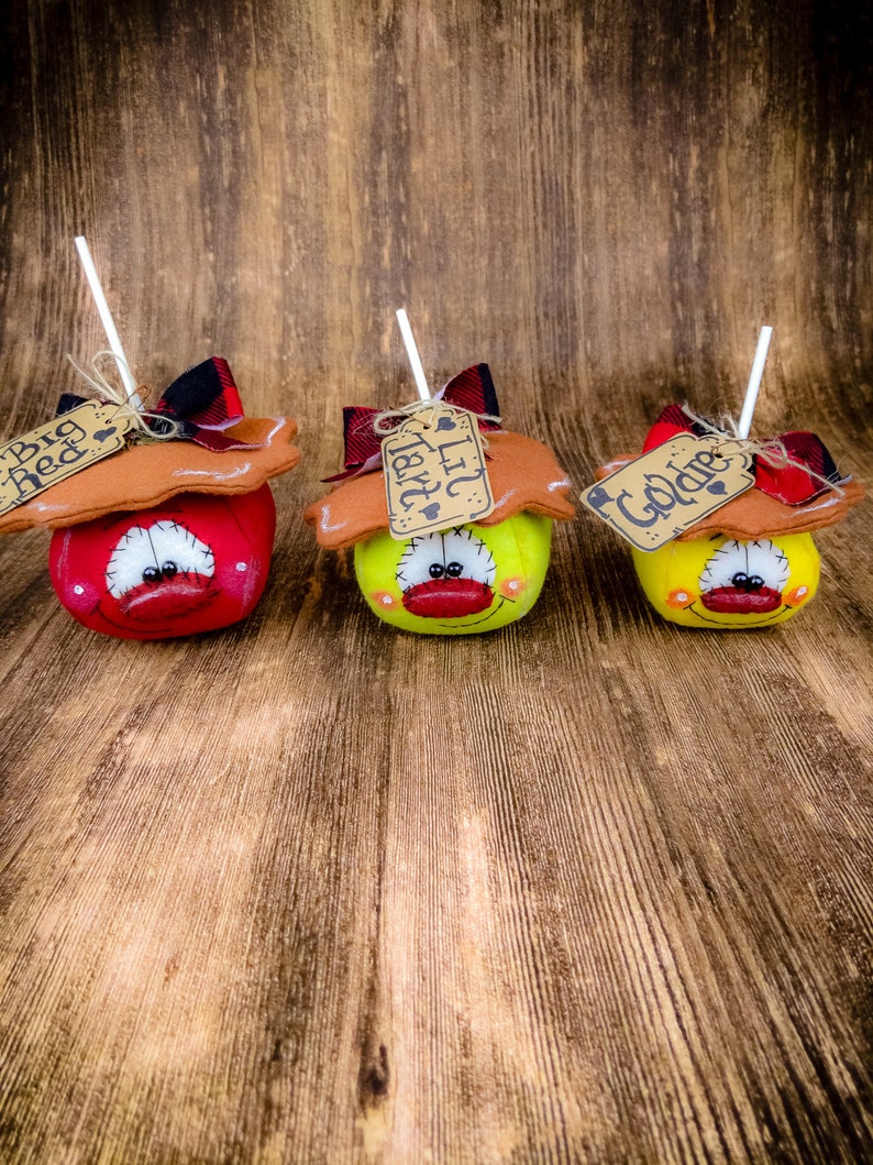 May include: Three handmade fabric apples with stick stems and brown paper tags. The apples are red, yellow, and green. The tags read "Big Red", "Lil'  Tater", and "Golden".