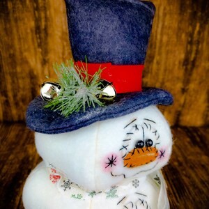 Pu&ograve; includere: Un pupazzo di neve bianco con un cappello di feltro blu, un nastro rosso e campanelli dorati. Il pupazzo di neve ha un viso cucito con occhi neri e un naso arancione. Il pupazzo di neve indossa una sciarpa bianca con un motivo a fiocco di neve.