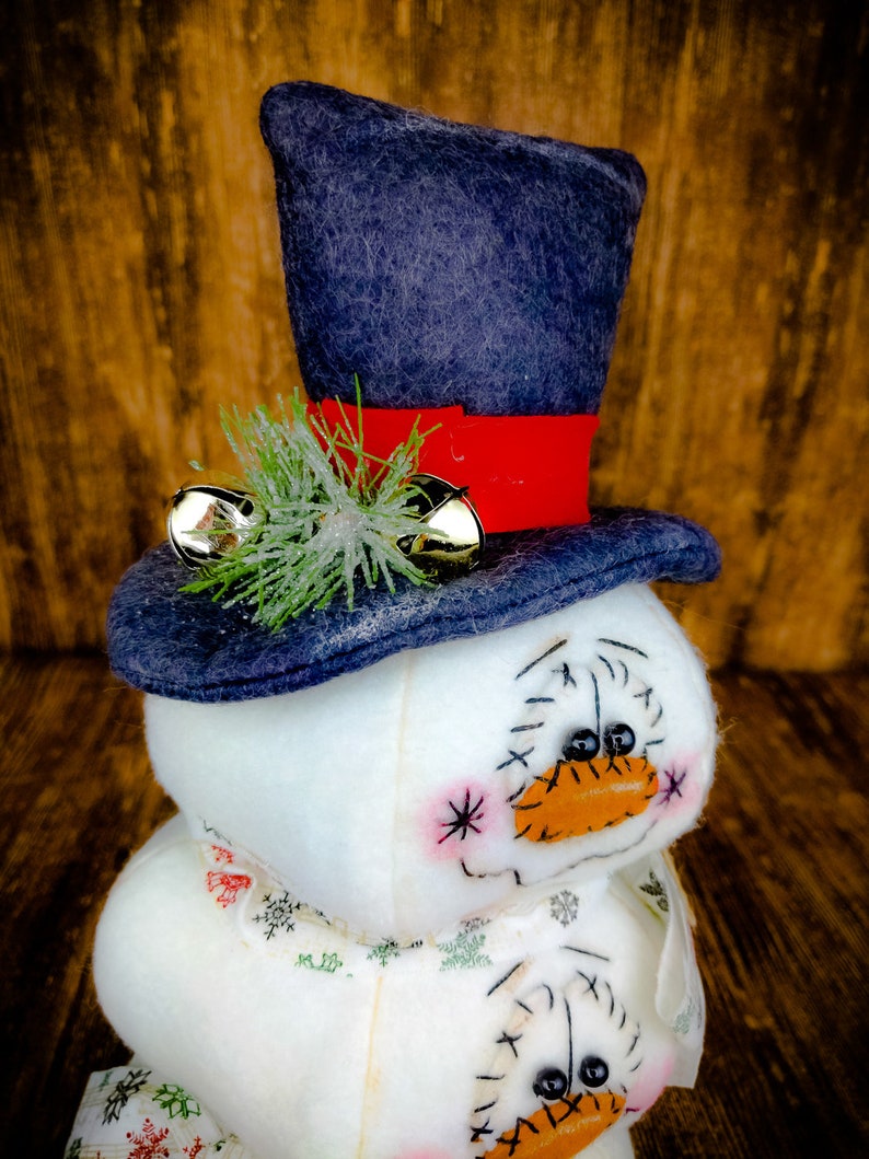 Pu&ograve; includere: Un pupazzo di neve bianco con un cappello di feltro blu, un nastro rosso e campanelli dorati. Il pupazzo di neve ha un viso cucito con occhi neri e un naso arancione. Il pupazzo di neve indossa una sciarpa bianca con un motivo a fiocco di neve.