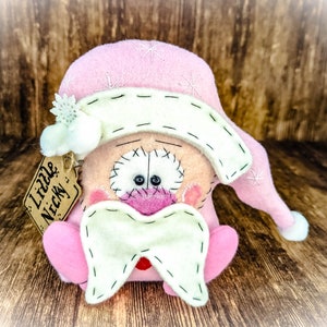 May include: A handcrafted felt doll featuring a pink hat with white embroidered snowflakes, a cream-coloured brim, and a matching cream beard. The doll has a round face with stitched details, pink cheeks, and a tag attached that reads Little Nicky.