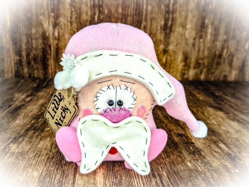 May include: A handcrafted felt doll featuring a pink hat with white embroidered snowflakes, a cream-coloured brim, and a matching cream beard. The doll has a round face with stitched details, pink cheeks, and a tag attached that reads Little Nicky.