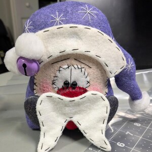 May include: A handcrafted felt gnome figurine featuring a purple hat with white embroidered snowflakes and a white pom-pom. The gnome has a white beard, black bead eyes, and a purple jingle bell. A ruler shows it is approximately 20 cm tall.