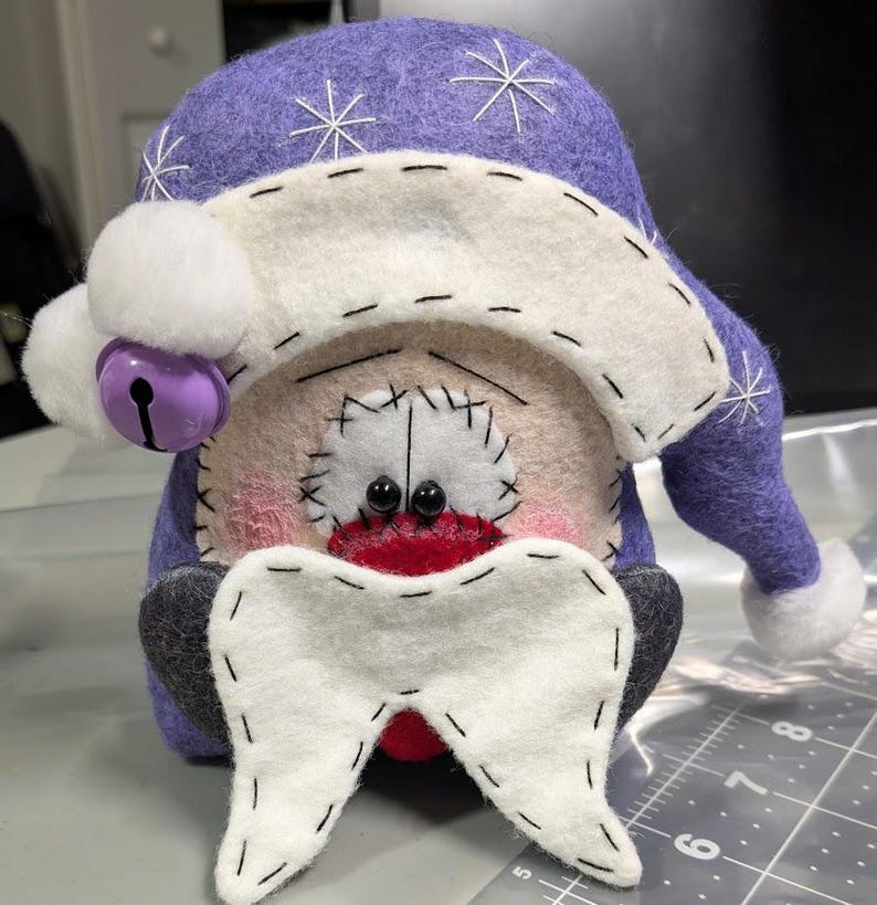 May include: A handcrafted felt gnome figurine featuring a purple hat with white embroidered snowflakes and a white pom-pom. The gnome has a white beard, black bead eyes, and a purple jingle bell. A ruler shows it is approximately 20 cm tall.