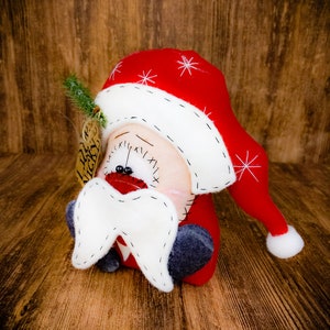 May include: A plush Santa Claus figure with a red hat featuring white embroidered snowflakes and a white pom-pom. The figure has a white felt moustache, black bead eyes, and a stitched mouth. A small brown tag labelled Lil Nicky is attached.