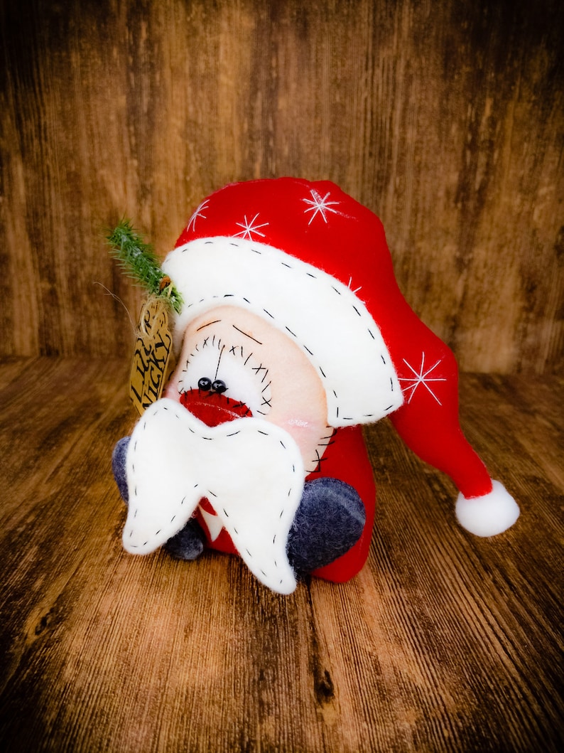 May include: A plush Santa Claus figure with a red hat featuring white embroidered snowflakes and a white pom-pom. The figure has a white felt moustache, black bead eyes, and a stitched mouth. A small brown tag labelled Lil Nicky is attached.