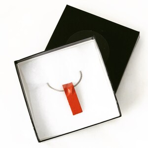 May include: An orange rectangular pendant necklace presented in an open black gift box with a white interior. The pendant hangs from a silver-coloured chain. The box is on a white background.