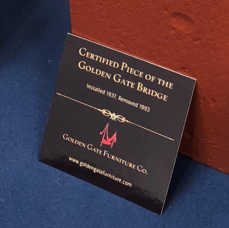 May include: A black card with gold lettering that reads "Certified Piece of the Golden Gate Bridge Installed 1937, Removed 1993". Below the text is a red logo of a bridge with the text "Golden Gate Furniture Co." and the website "www.goldengatefurniture.com".