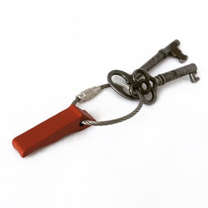 May include: Two antique skeleton keys linked by a steel cable and a red rectangular key fob. The keys are dark grey with ornate designs. The key fob is a solid, muted red colour.