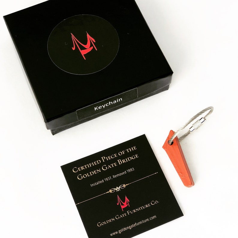 May include: A black box with a red logo and the word "Keychain" on a label. A small, orange, triangular keychain with a silver wire loop is next to the box. A black card reads "Certified Piece of the Golden Gate Bridge."