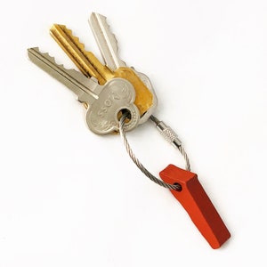 May include: A set of keys attached to a silver cable keychain with a red tag. The keys are silver and gold-toned. One key has the word "MOSS" on it. The keychain is against a white backdrop.