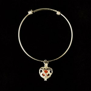 May include: A silver bangle bracelet with a heart-shaped charm. The charm is open and has a delicate design. The bracelet is adjustable.