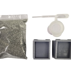 May include: A clear plastic bag filled with small, dark green granules, a clear plastic dropper with a bulb, and two gray, square containers with brick-like detailing. The items are likely for a craft or hobby project.