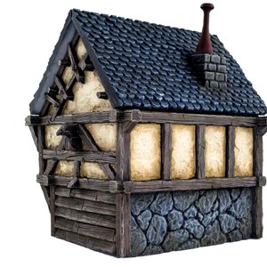 May include: A miniature wooden house with a blue roof, a chimney, and a stone foundation. The house has a half-timbered facade with exposed wooden beams and a weathered, cream-colored plaster.