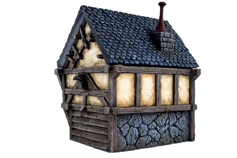 May include: A miniature wooden house with a blue roof, a chimney, and a stone foundation. The house has a half-timbered facade with exposed wooden beams and a weathered, cream-colored plaster.