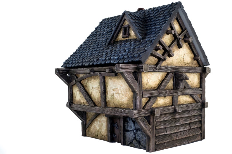 May include: A miniature wooden house with a dark blue shingled roof, a small window, and a stone foundation. The house has a half-timbered facade with exposed wooden beams and a weathered, cream-colored plaster.