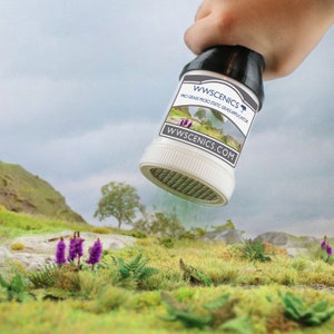 May include: A white plastic container with a black lid and a green mesh screen. The container is labeled "WWScenics Pro Grass Micro Static Grass Applicator WWScenics.com". The container is being held over a green grassy surface with purple flowers.
