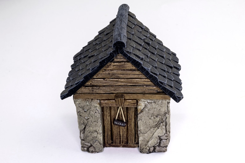 May include: A small, wooden building with a black shingled roof and a stone foundation. The door is wooden and has a sign that says "Vacant".