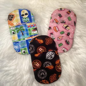 May include: Three different fabric pouches with various designs. One pouch has a tropical design with a pineapple, palm trees, and a checkered pattern. Another pouch has a pink background with a sushi design. The third pouch has a black background with a chicken wing design and the text "I'm just here for the wings".