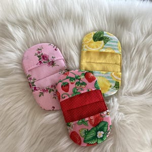 May include: Three fabric oven mitts with colorful patterns. One is pink with floral designs, another features strawberries, and the third has a lemon theme. The mitts are arranged on a white, fluffy surface.