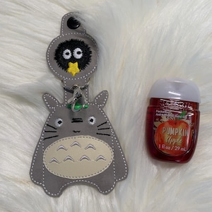 May include: A gray and beige Totoro keychain with a black and yellow Totoro charm attached. The keychain is next to a 1 fl oz / 29 ml bottle of hand sanitizer with a red label that says "Pumpkin Apple".