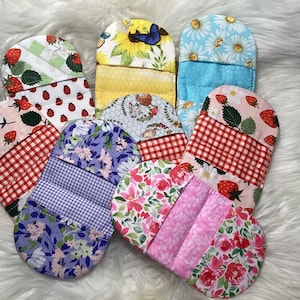 May include: A collection of seven rounded fabric pot holders featuring various patterns including strawberries, ladybugs, sunflowers with a butterfly, daisies, and floral prints in shades of red, yellow, blue, purple, pink, and green with gingham accents.