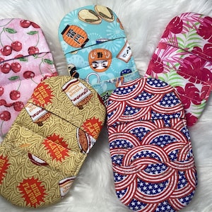 May include: Five colorful pot holders with different patterns. The patterns include cherries, ramen noodles, American flags, and tropical leaves.
