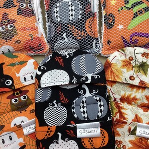 May include: A collection of rectangular kitchen scrubbers and rounded pot holders featuring various seasonal patterns including owls, pumpkins, ghosts, and autumn leaves. Each item has a white tag with the text Sanity printed in black.