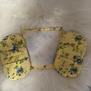 May include: A set of three kitchen items: two oven mitts and a square hot pad. The items are yellow with a floral pattern of blue flowers and green leaves. The hot pad has a white grid pattern.