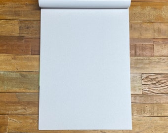 May include: A vertical rectangular white notepad with a thick white binding at the top edge. The paper has a subtle textured surface and is oriented in a portrait position, featuring a clean, blank white space for writing or drawing purposes.