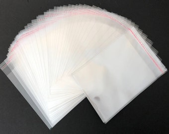 May include: A stack of clear, rectangular plastic bags arranged in a fan shape. Each bag features a thin, red adhesive strip along the top edge for sealing. The bags are made of a transparent, flexible material and are stacked neatly together.