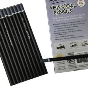 May include: A set of twelve black charcoal pencils with grey end caps, arranged in a row next to their light purple packaging. The box features the text "CHARCOAL PENCILS" and "12pcs Mont Marte Charcoal Pencils 3 Size Set" with an 8mm round body.