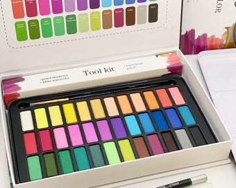 May include: A watercolor paint set in a white box featuring a color chart on the lid, containing thirty-six rectangular paint pans, a black 2B pencil, a clear water brush, a metal pencil sharpener, and a small tube of white watercolor paint.