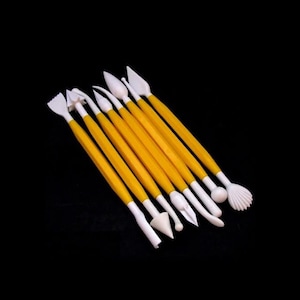 Op de afbeelding: Een set van acht dubbelzijdige boetseerinstrumenten met felgele handvatten en witte plastic uiteinden. Elk instrument heeft uiteinden met verschillende vormen, waaronder punten, bollen, schelpen en gekartelde randen voor het vormen en bewerken.