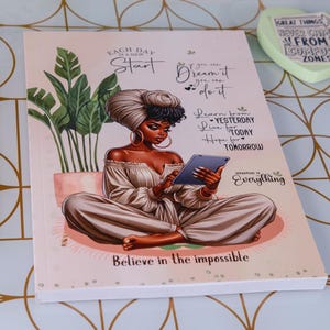 May include: A notebook featuring an illustration of a woman with a tablet, with the words "Believe in the impossible." A gold pen and two pastel highlighters are on the side. A heart-shaped decoration is also visible.