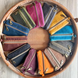 Puede incluir: Una colección de monederos con cremallera de colores, dispuestos en círculo sobre una bandeja de madera. Los monederos vienen en varios colores, incluyendo azul, verde, rosa, amarillo y naranja. Cada monedero tiene una cremallera metálica.